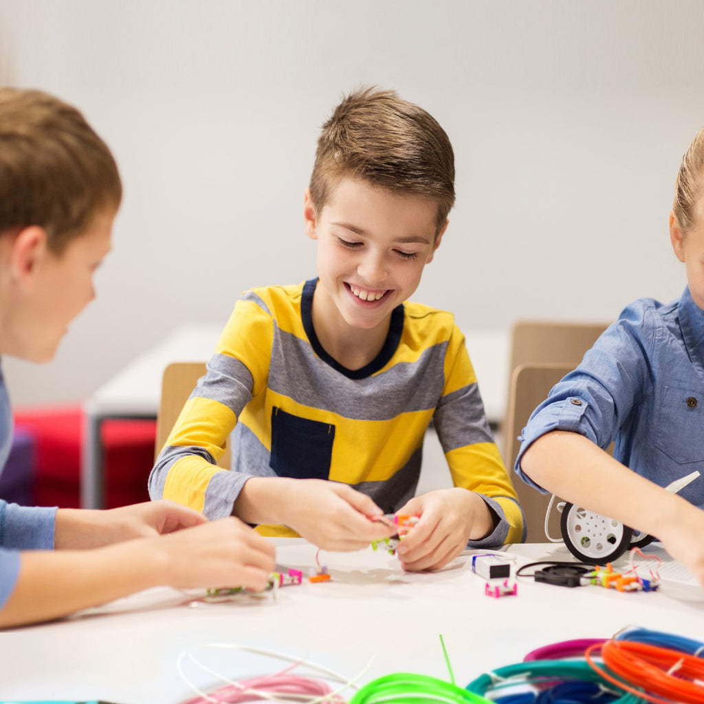 Three children smiling and engaged in a 3D pen project with colorful filament spools on a table.