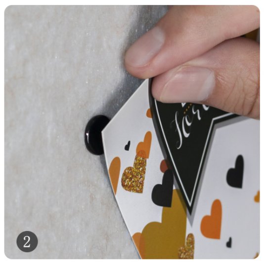 Hand holding a card with hearts, attaching it to a magnetic pushpin on a textured surface. Number 2 overlay.