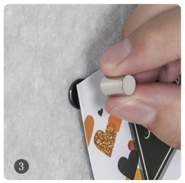 Hand holding a cylindrical magnet to attach a card with hearts to a textured surface with a black pushpin.