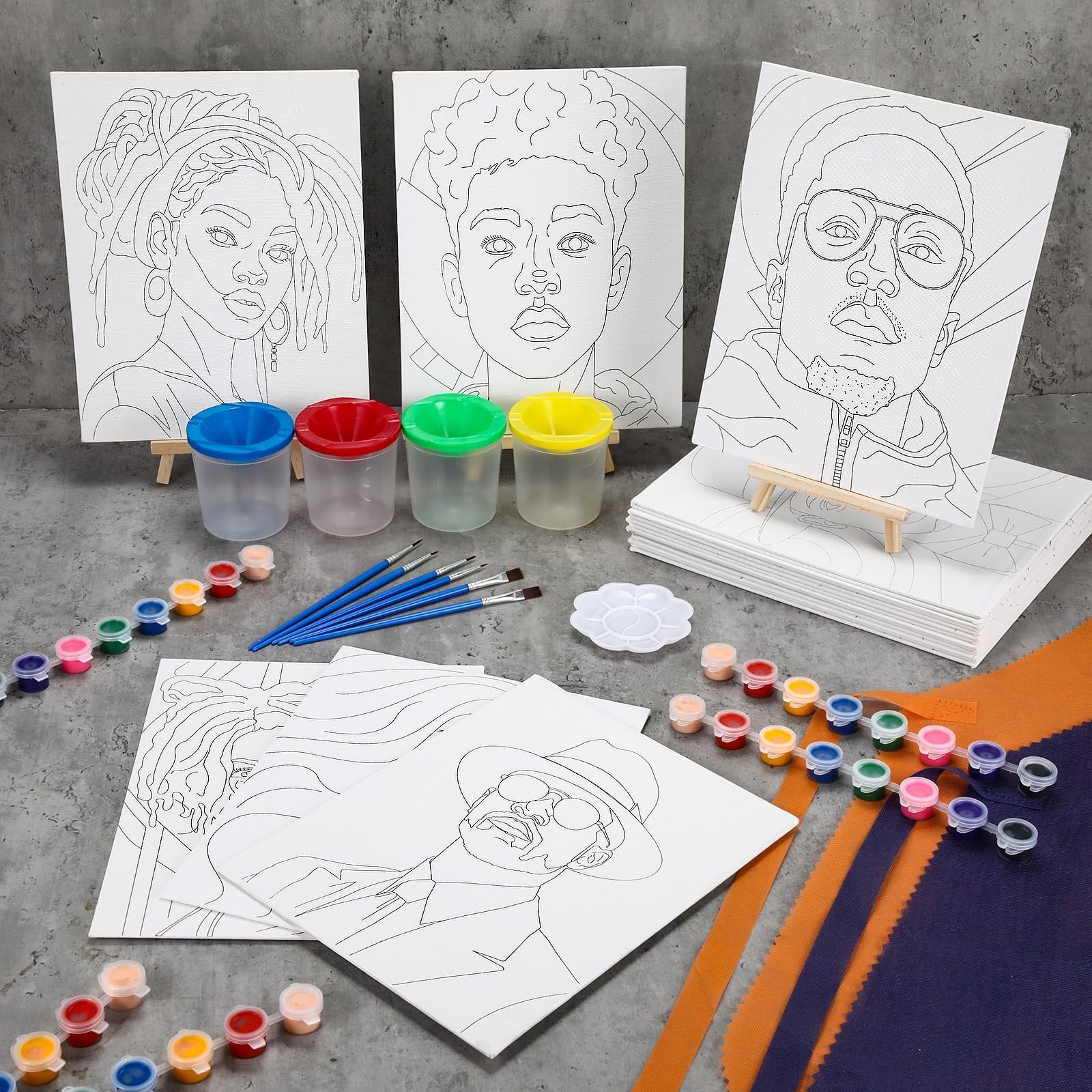 Pre-drawn canvas painting kit with 3 canvases featuring portraits, paint pots, brushes, and aprons laid out on a gray surface.