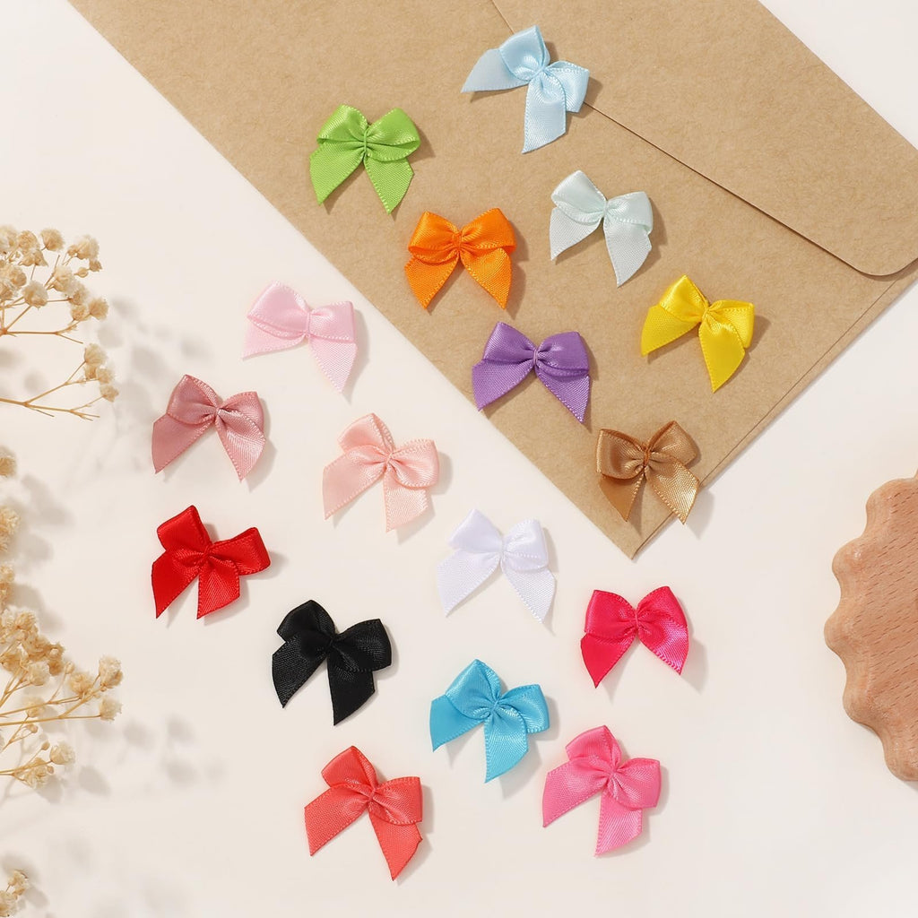 Assortment of 1-inch mini ribbon bows in various colors scattered on a light surface with a brown envelope.