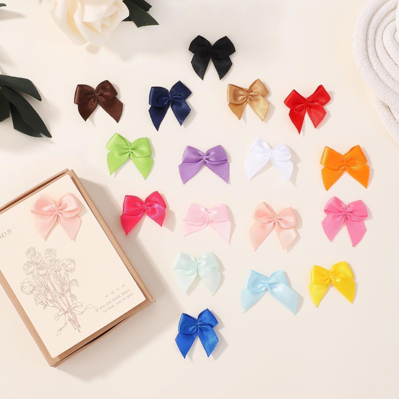 Assortment of 1-inch mini ribbon bows in various colors scattered on a light surface, with a decorative card on the left.