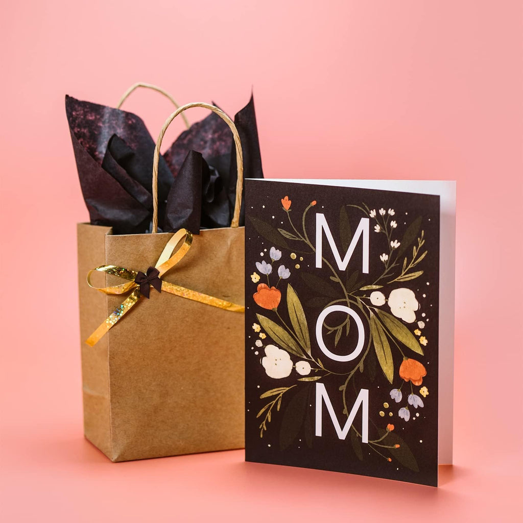 Gift bag with tissue paper and a card that says MOM, set against a pink background.