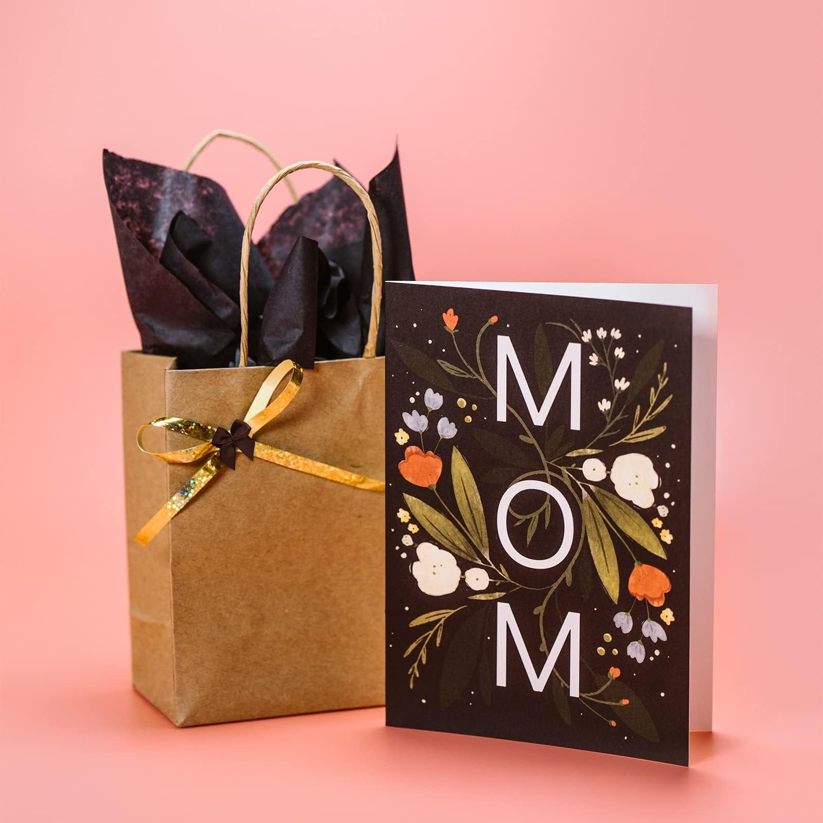 Gift bag with tissue paper and a card that says MOM, set against a pink background.