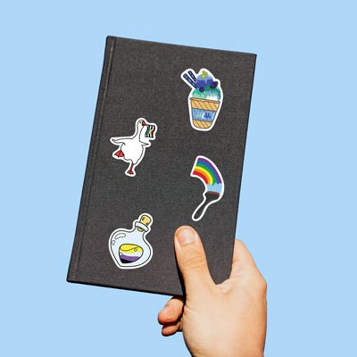 Hand holding a black notebook with four LGBTQ+ pride stickers: a goose with a rainbow flag, a rainbow paintbrush, a potion bottle with nonbinary flag colors, and a cup of blue dessert.