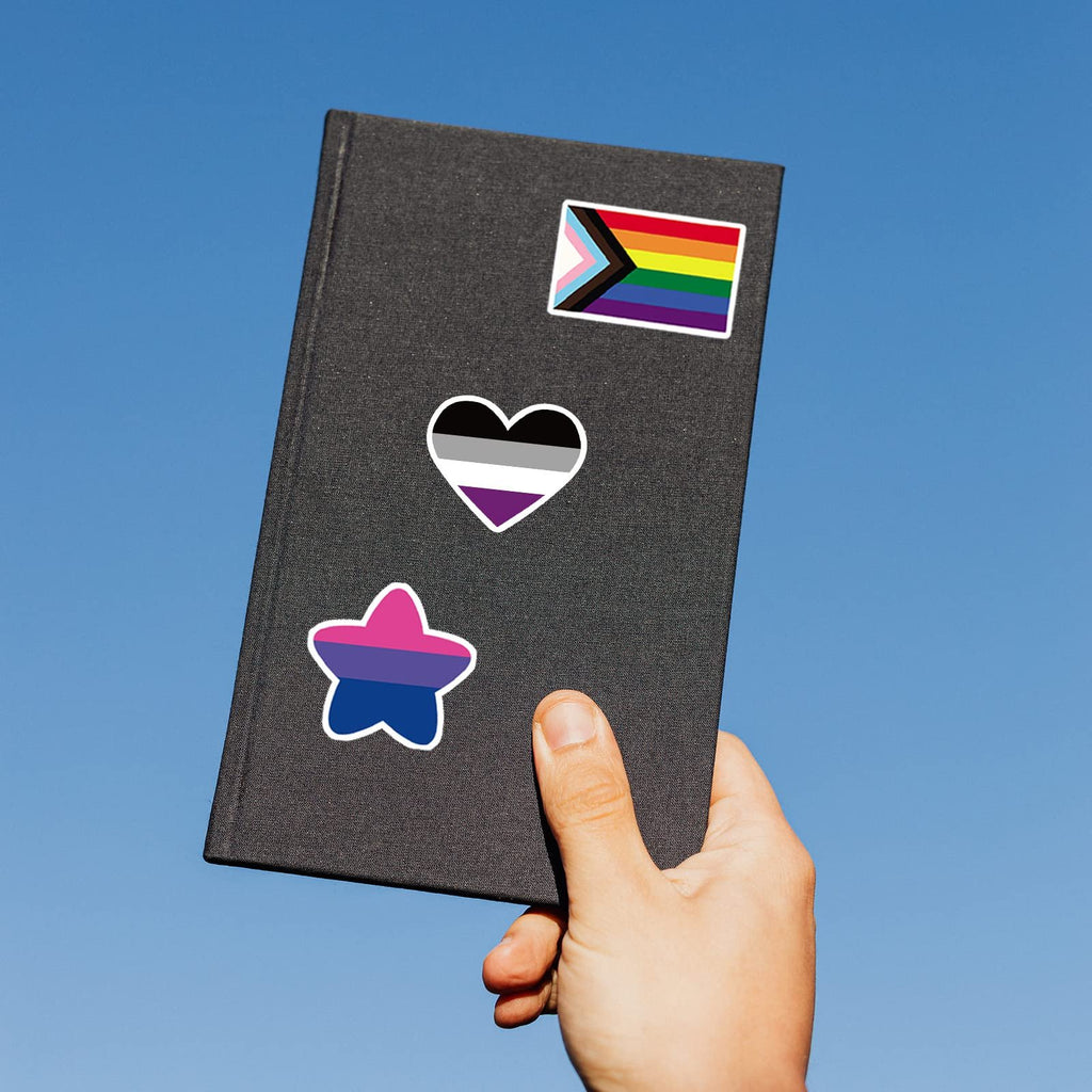 Hand holding a dark gray notebook with three LGBTQ+ pride flag stickers: a progress pride flag, an asexual pride flag heart, and a bisexual pride flag star.