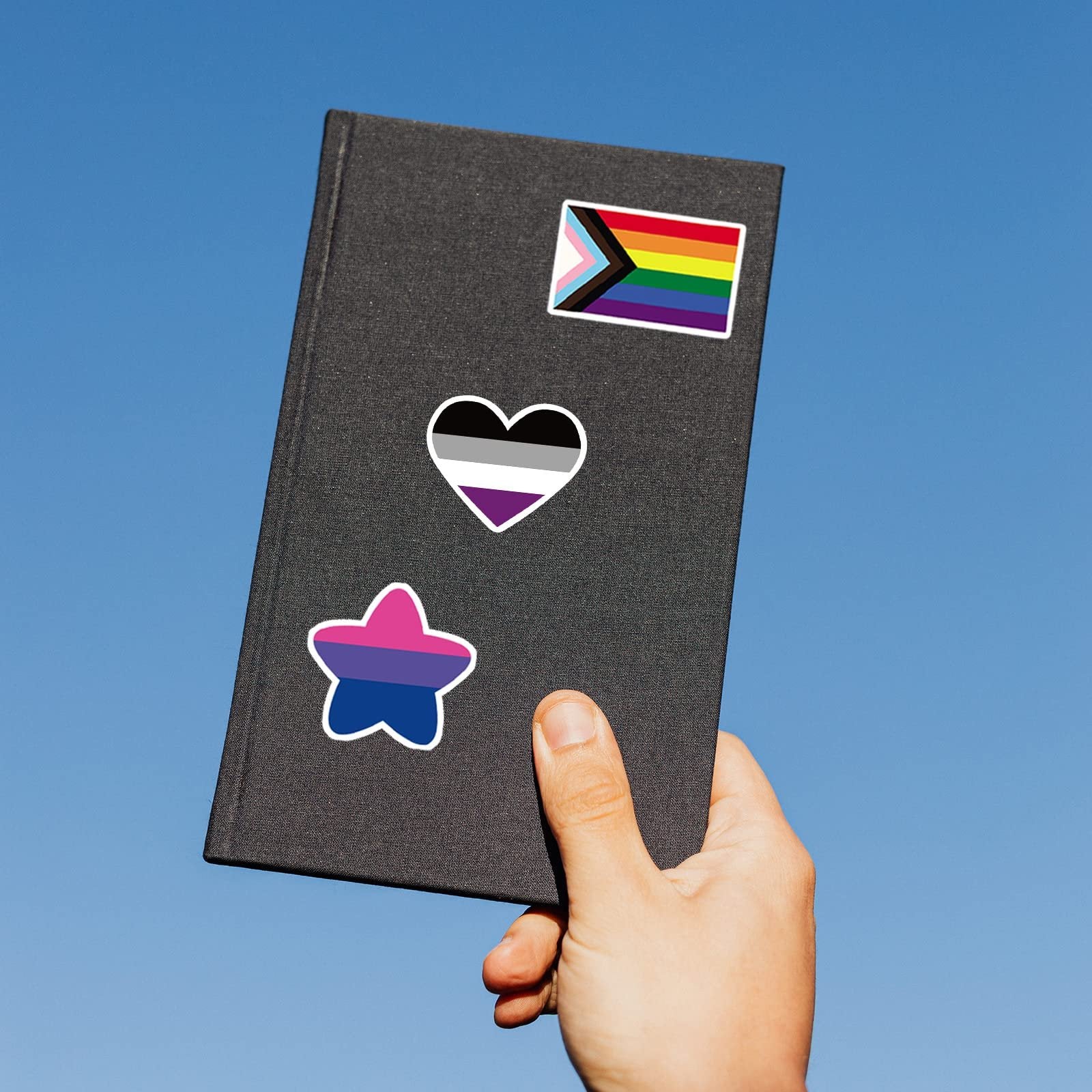 Hand holding a dark gray notebook with three LGBTQ+ pride flag stickers: a progress pride flag, an asexual pride flag heart, and a bisexual pride flag star.