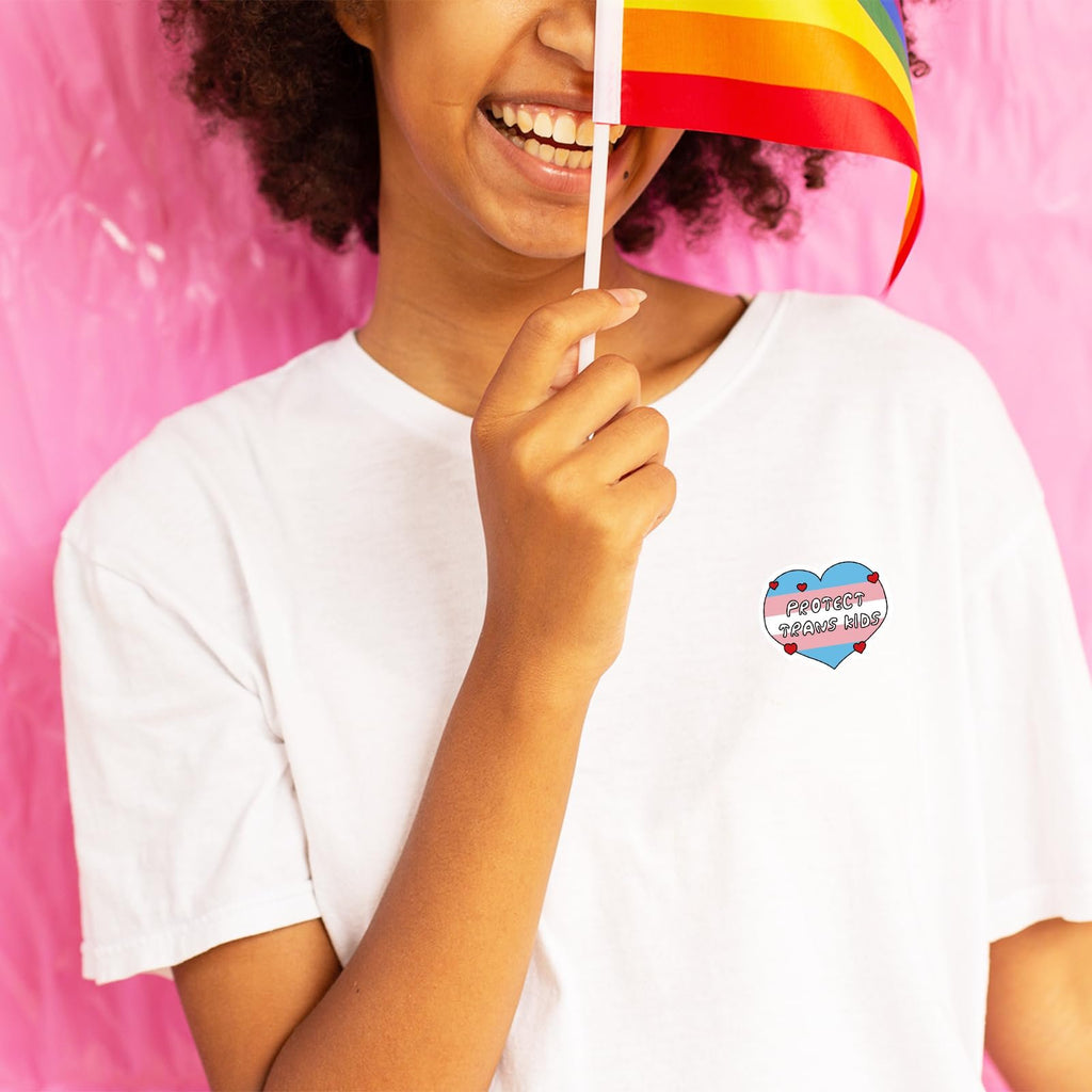 Person wearing a white t-shirt with a heart-shaped sticker that says 'PROTECT TRANS KIDS' and holding a rainbow flag.