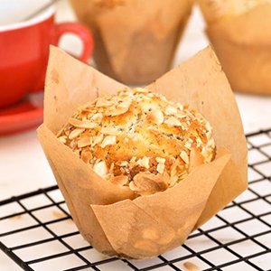 Almond muffin in a natural tulip cupcake liner on a cooling rack, with a red mug in the background.