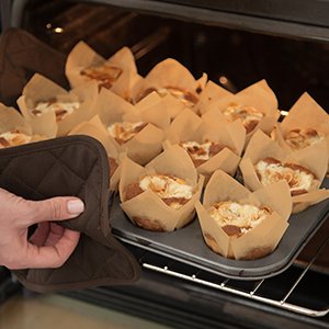Hand wearing oven mitt holding muffin tin with tulip cupcake liners filled with baked muffins being removed from oven