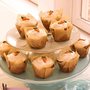Carrot cake cupcakes with white frosting and orange carrot decorations, presented on a two-tier cake stand.