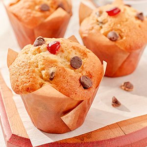 Three chocolate chip and cherry muffins in tulip-shaped parchment paper liners on a wooden board.