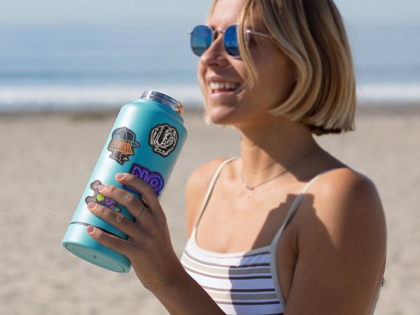 Woman holding a teal water bottle with various stickers on a beach