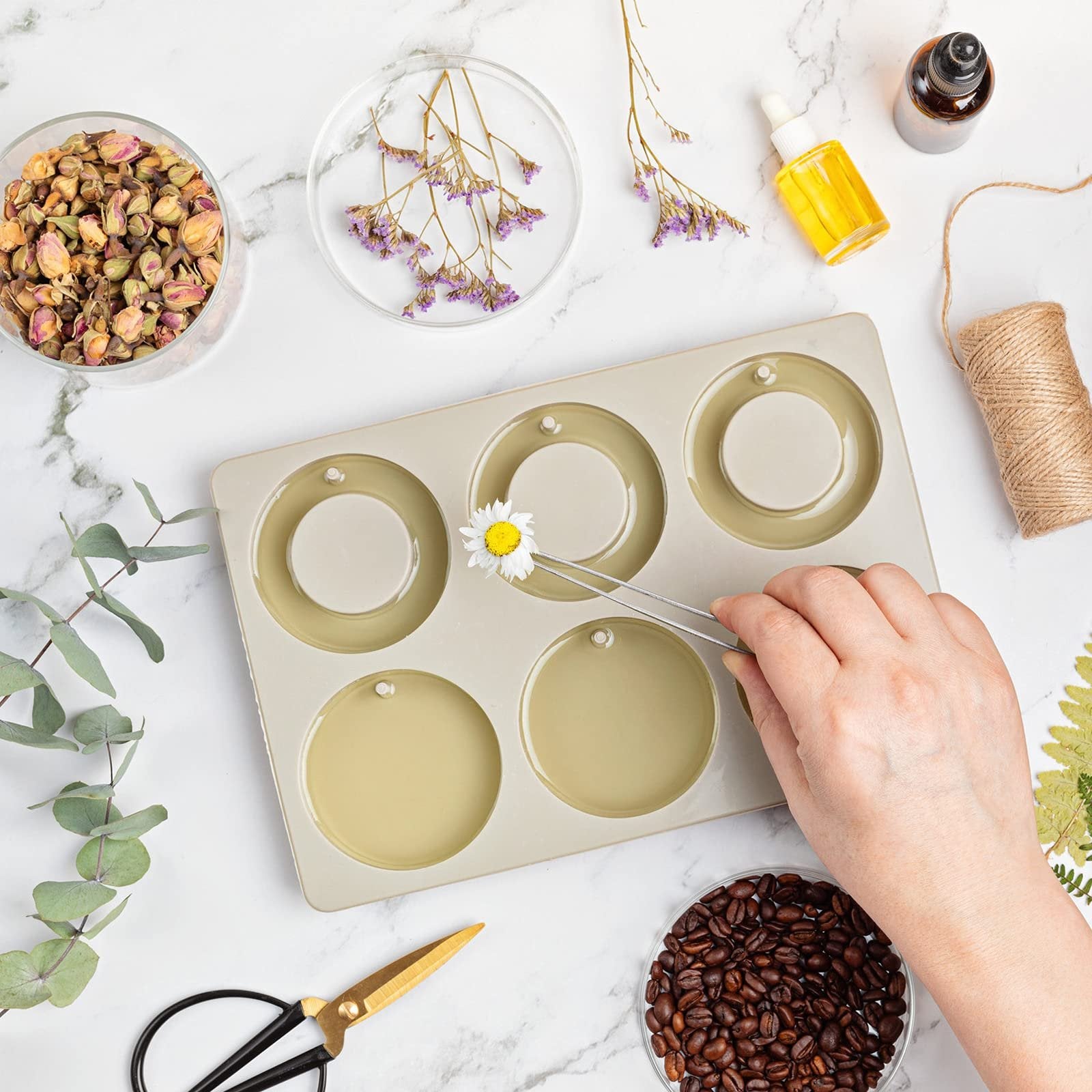 Hand using tweezers to place a daisy into a round silicone mold for crafts, surrounded by dried flowers, coffee beans, and crafting supplies.