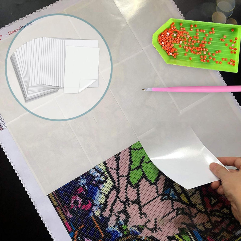 Diamond painting canvas with a hand peeling back a white release paper sheet, showing a stack of papers and a tray of diamonds.