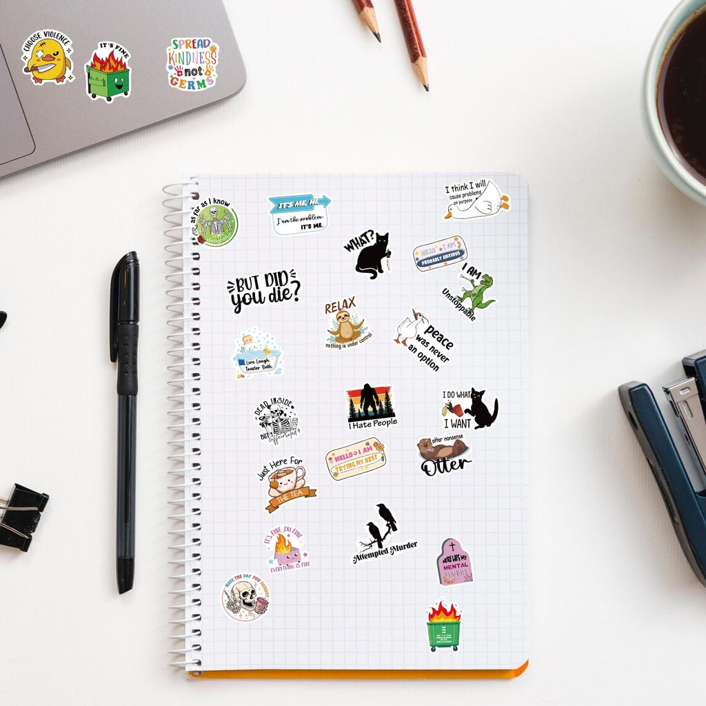 Assortment of funny vinyl stickers scattered on a notebook page and laptop, featuring various designs and text.