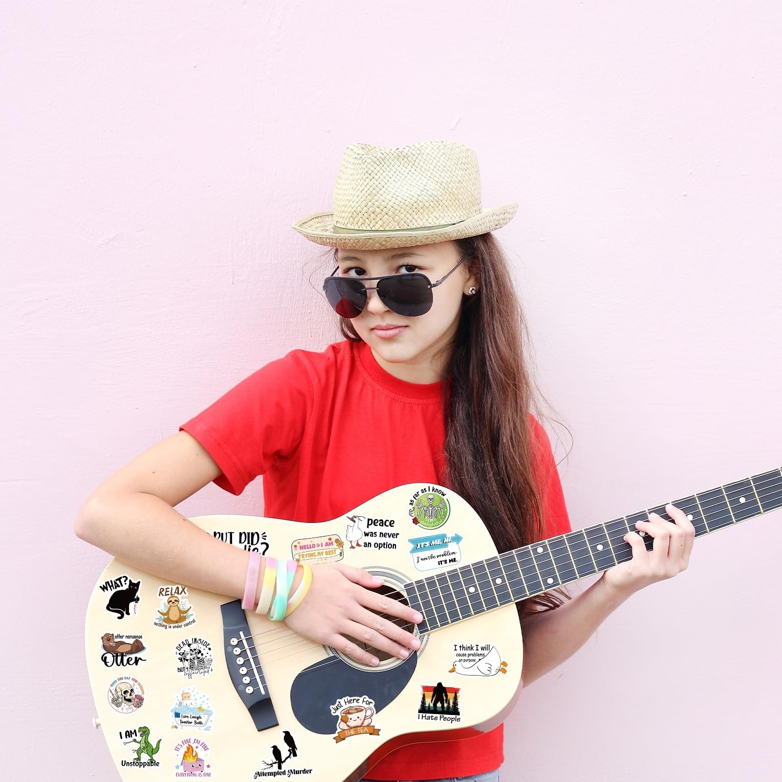 Young person wearing a hat and sunglasses plays a guitar decorated with various funny stickers.