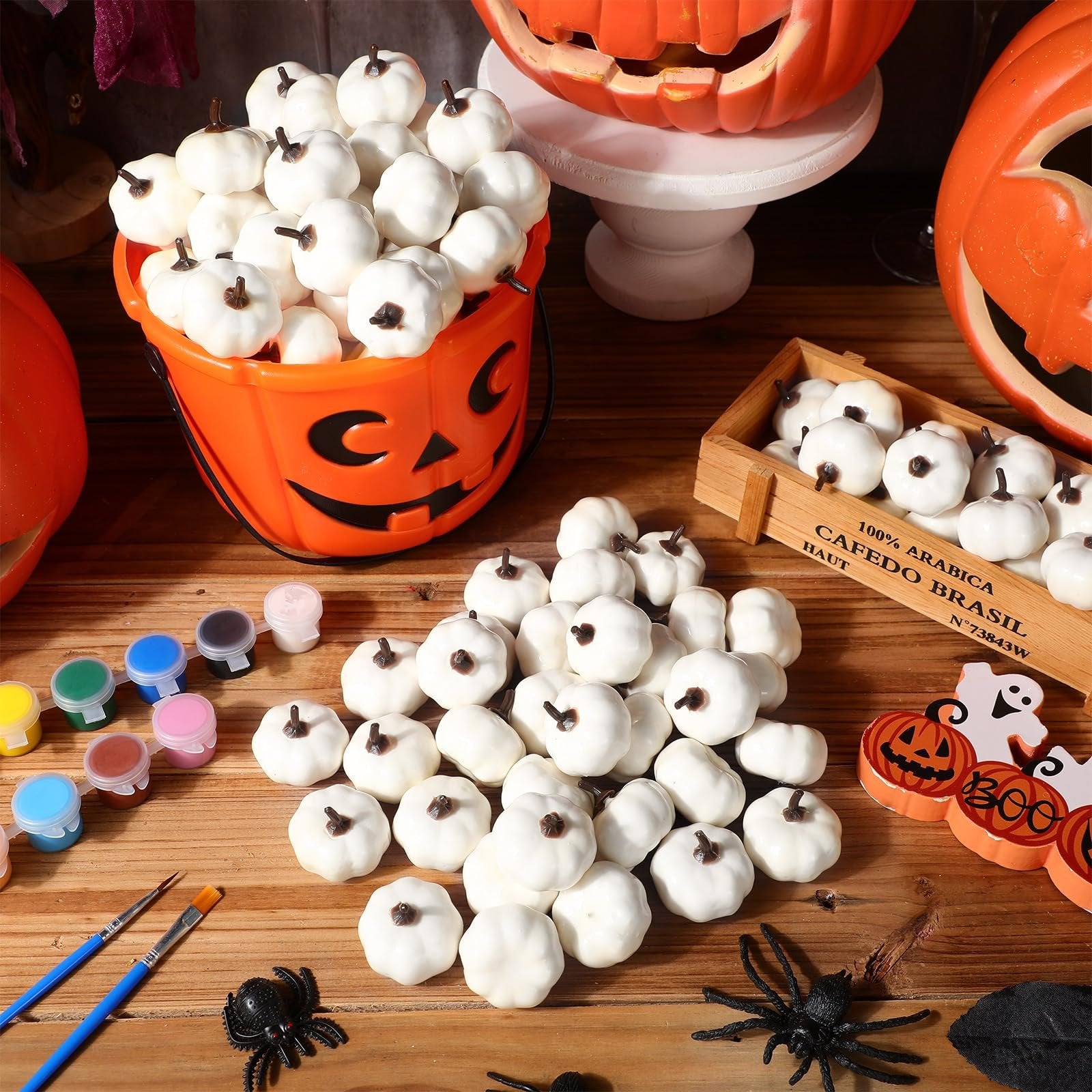 Mini white foam pumpkins with paint and brushes scattered on a wooden surface, with a jack-o'-lantern bucket and wooden crate.