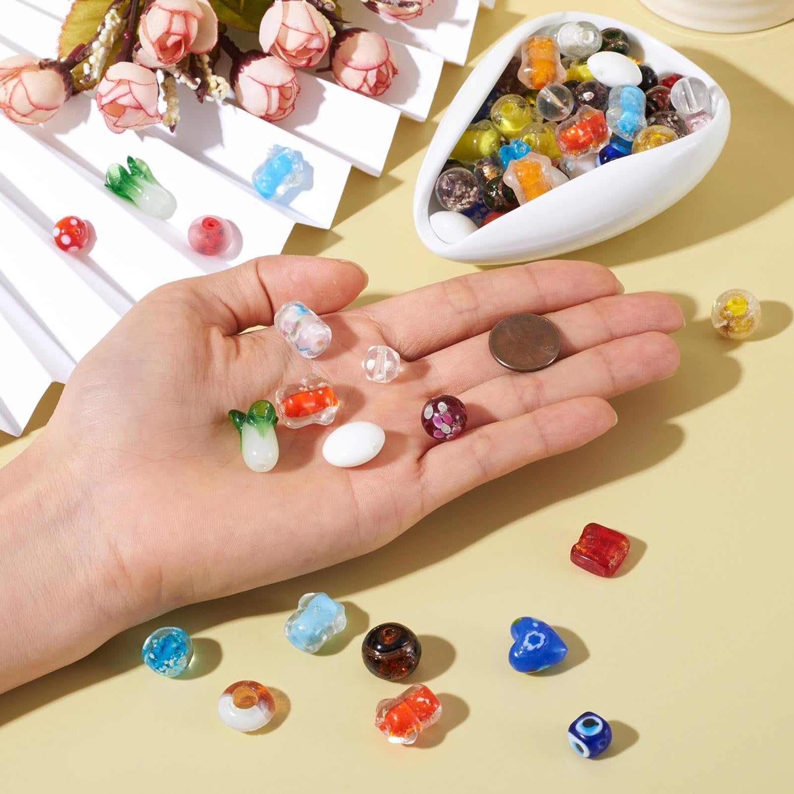 Hand holding assorted colorful lampwork glass beads, with a bowl of beads and decorative flowers in the background.