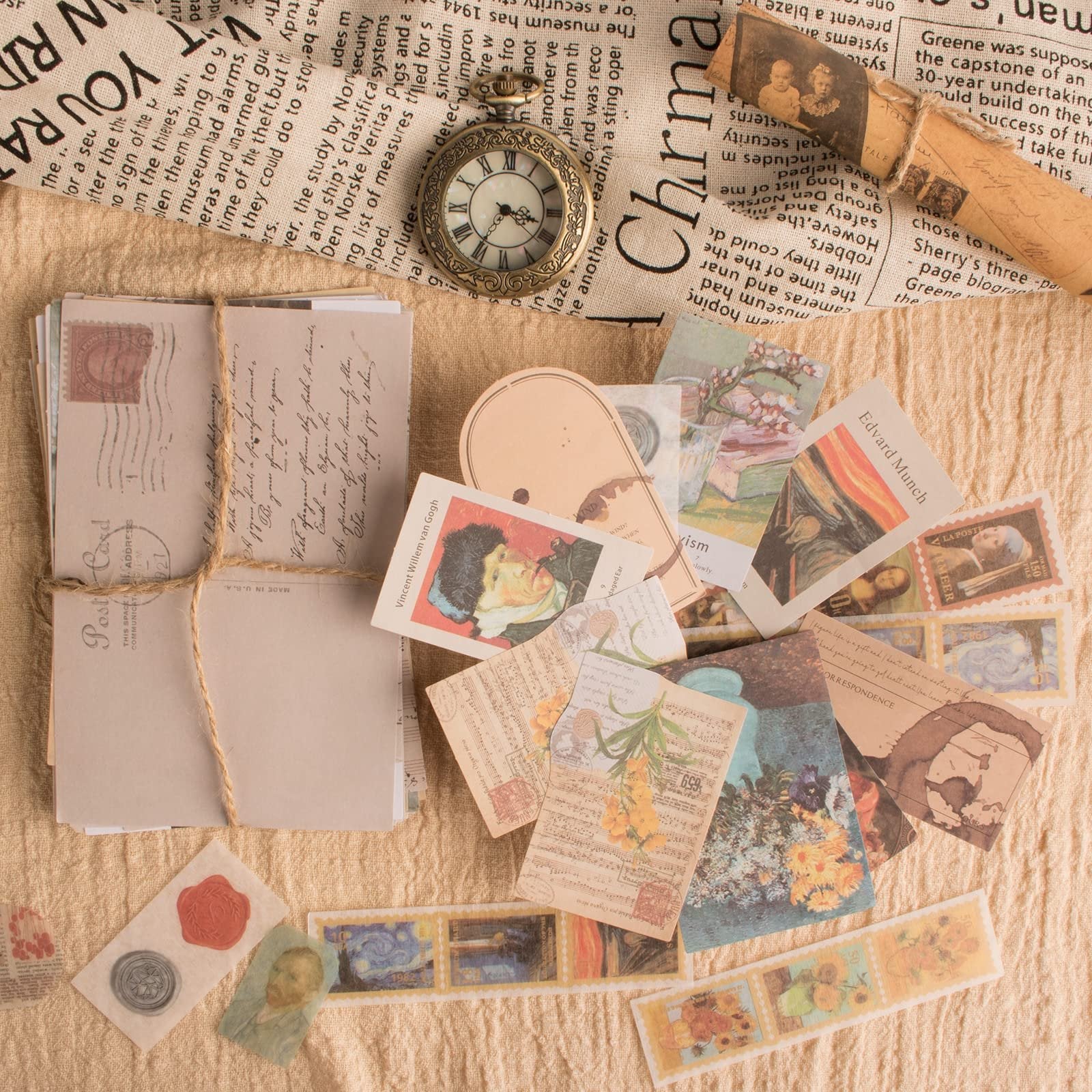 A flat lay of vintage scrapbook stickers, ephemera, and postcards scattered on a textured fabric background, with a pocket watch and wax seals.