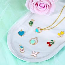 Assortment of colorful enamel and gold-tone jewelry charms on a white tray with a pink rose and green fabric backdrop.