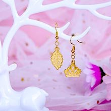 Two gold-tone dangle earrings, one shaped like a leaf and the other a crown, displayed on a white deer antler stand against a pink sparkly background.