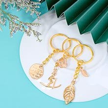 Three gold-tone keychain charms with decorative pendants including a leaf, a skier, and a coin-like disc, arranged on a white plate.