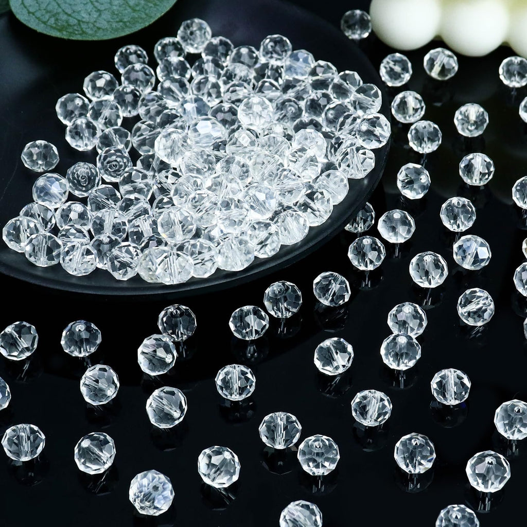 Pile of clear faceted crystal glass rondelle beads on a black plate and scattered on a black surface, with a green leaf and white spheres in the background.