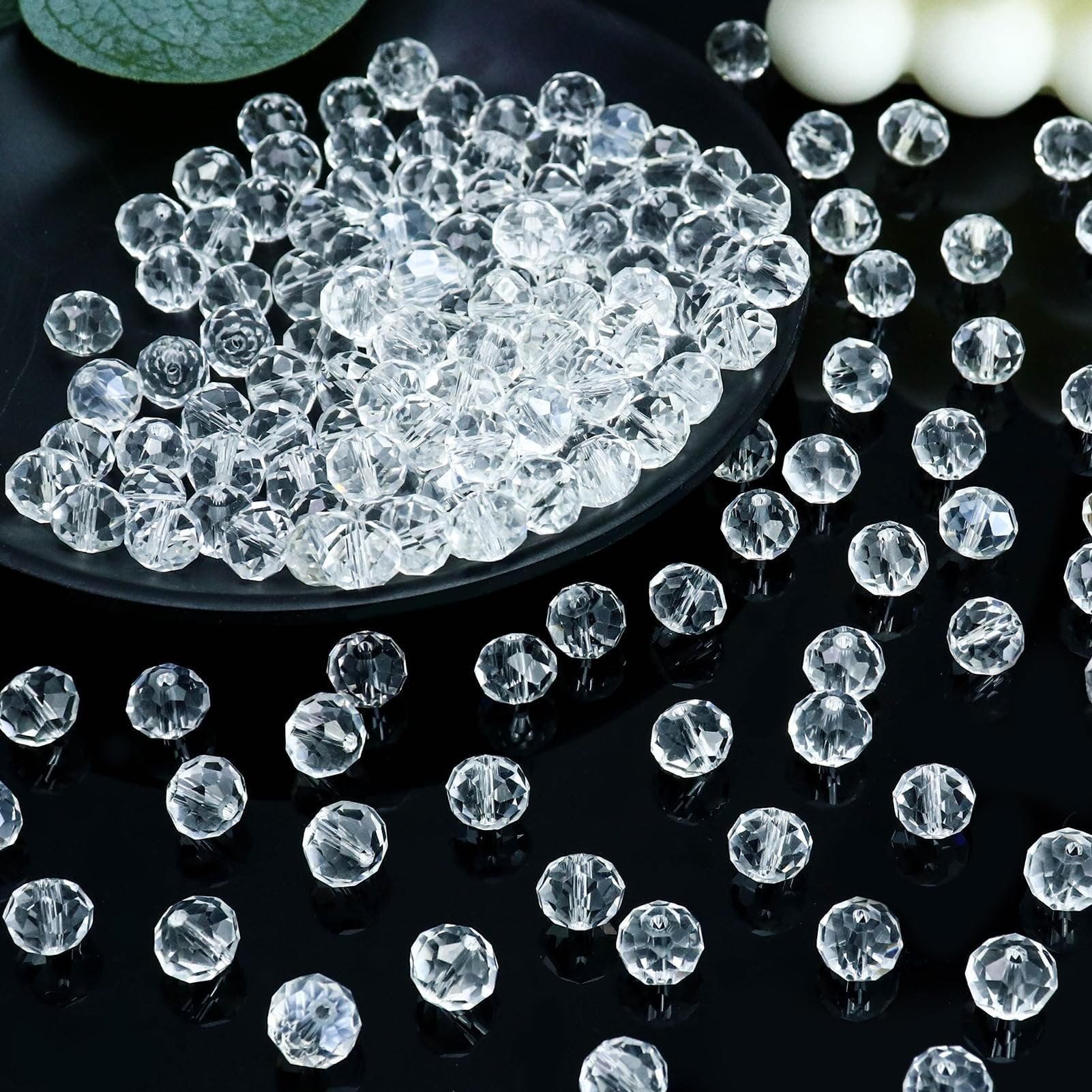 Pile of clear faceted crystal glass rondelle beads on a black plate and scattered on a black surface, with a green leaf and white spheres in the background.