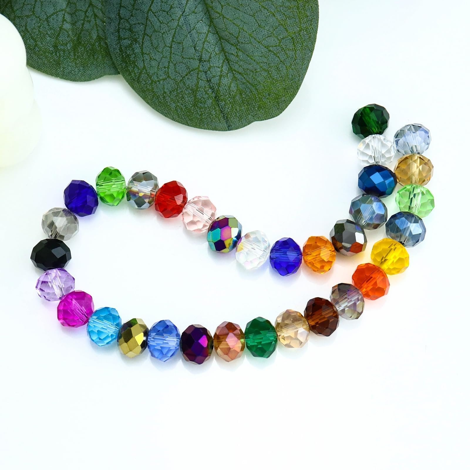 Assortment of colorful faceted crystal glass beads arranged in a curve on a white surface with a green leaf and white object in the background.