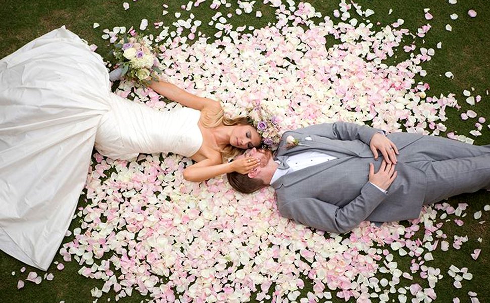 Bride and groom lying down on a bed of pink and white rose petals on grass, holding a bouquet.