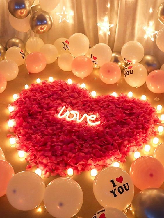 Heart-shaped arrangement of pink rose petals surrounded by LED tea lights and balloons, with a neon 'love' sign.