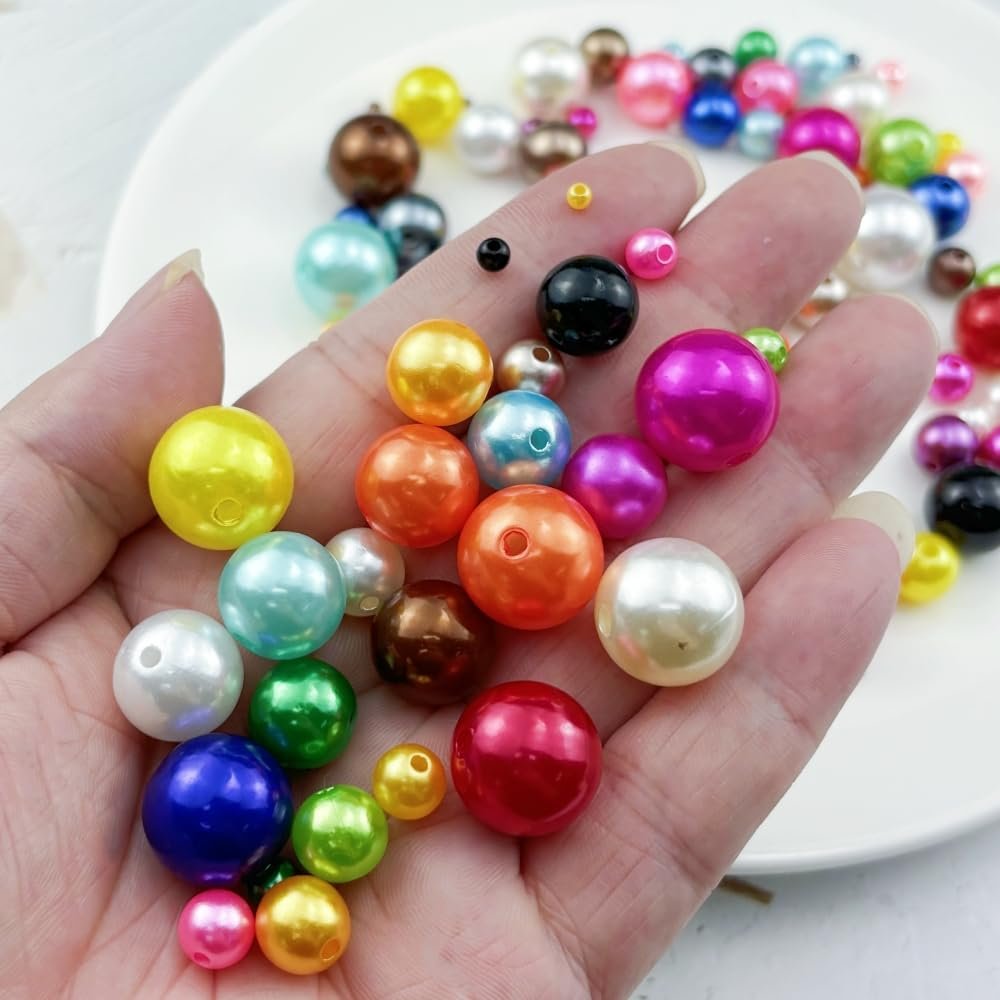 Hand holding a variety of colorful resin pearl beads for jewelry making, with more beads scattered on a white plate in the background.