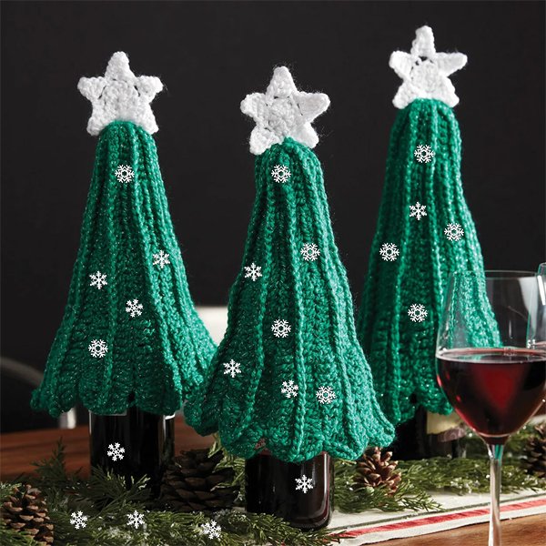 Three green knitted Christmas tree bottle covers with white stars on top, decorated with snowflakes, on a table with pinecones and a glass of red wine.
