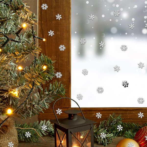 Mini metal snowflakes scattered on a wooden surface with a Christmas tree, lantern, and ornaments, viewed through a window with falling snow effect.