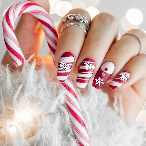 Close-up of a hand with festive red and white striped nails adorned with white snowflake charms, holding a candy cane.