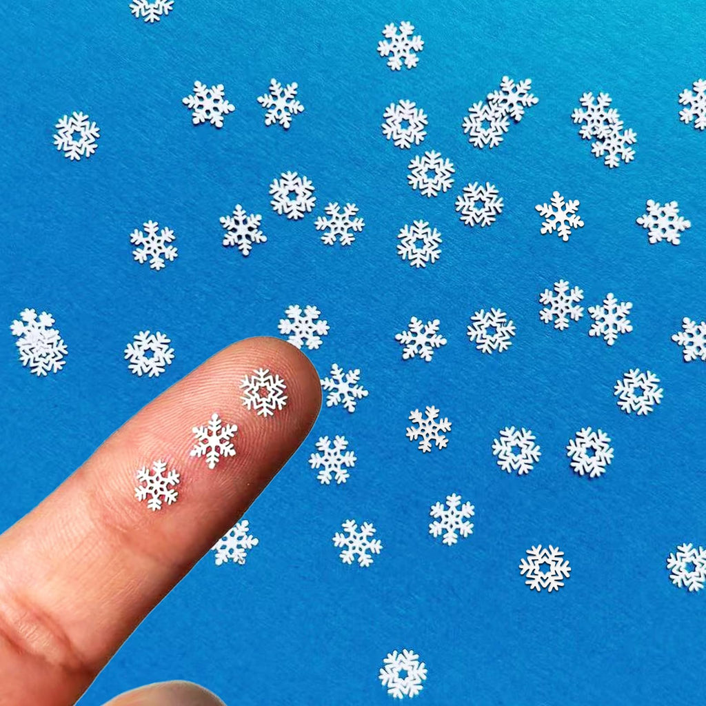 Close-up of a finger holding three small white metal snowflakes against a blue background with scattered snowflakes.