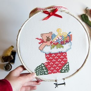 Hand holding embroidery hoop with a Christmas stocking cross-stitch pattern, red ribbon bow at top