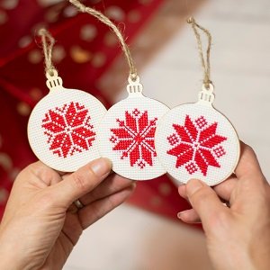 Three round wooden ornaments with red and white snowflake cross-stitch patterns, held by hands.