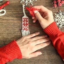 Hands in red sweater creating a woven ornament with red and white thread on a wooden surface with snowflakes