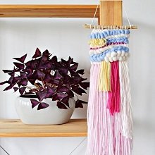 Woven wall hanging with pink, yellow, and blue yarn strands displayed on a shelf next to a potted plant.