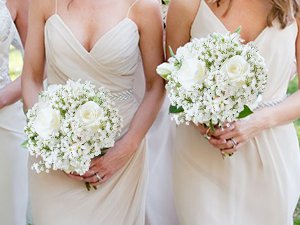 Two bridesmaids in beige dresses holding bouquets of white roses and baby's breath flowers.