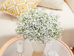 Bouquet of artificial baby's breath flowers in a white vase on a coffee table, with a candle and pillow in the background.