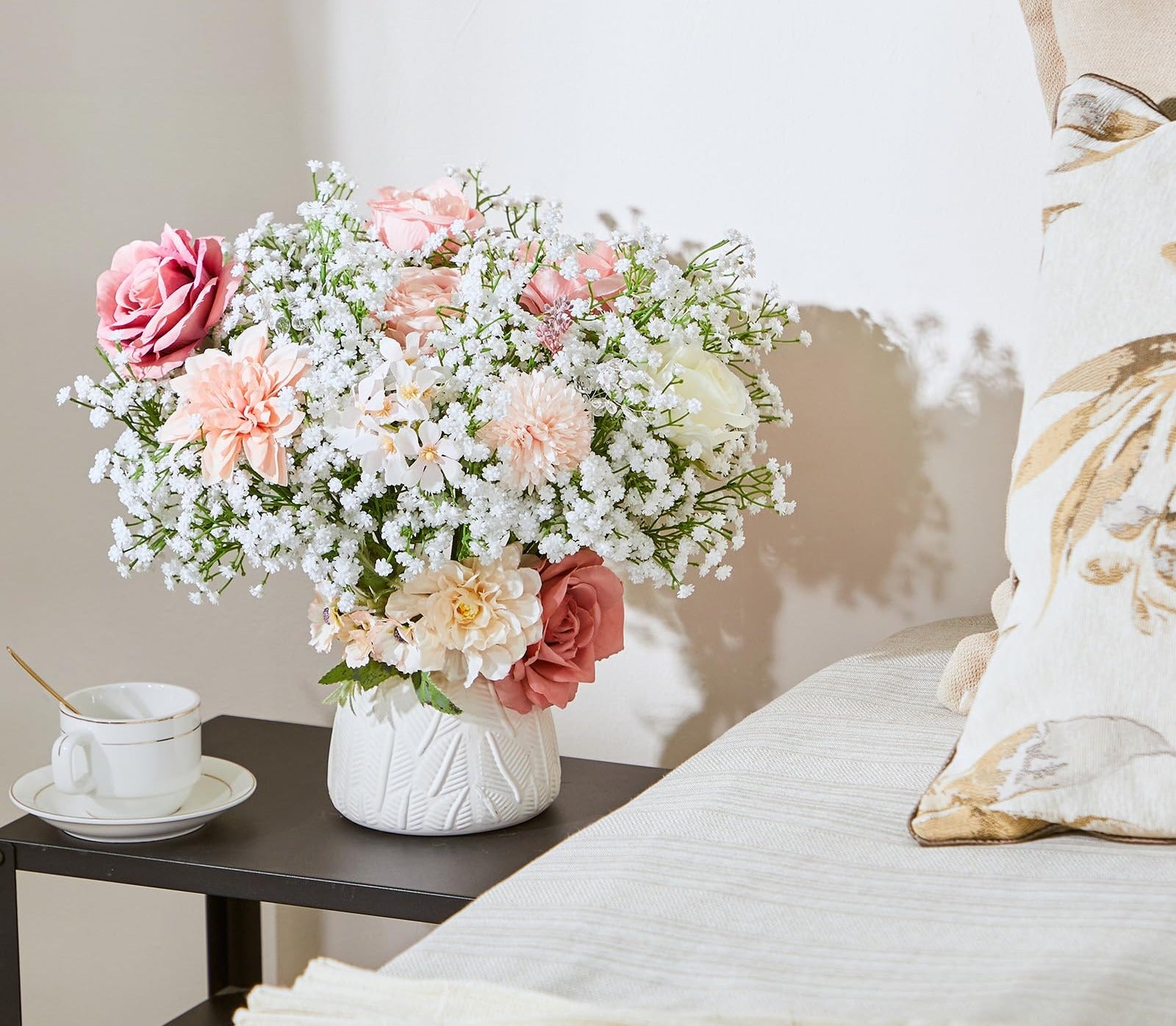 Artificial baby's breath and rose bouquet in a white vase on a side table with a teacup