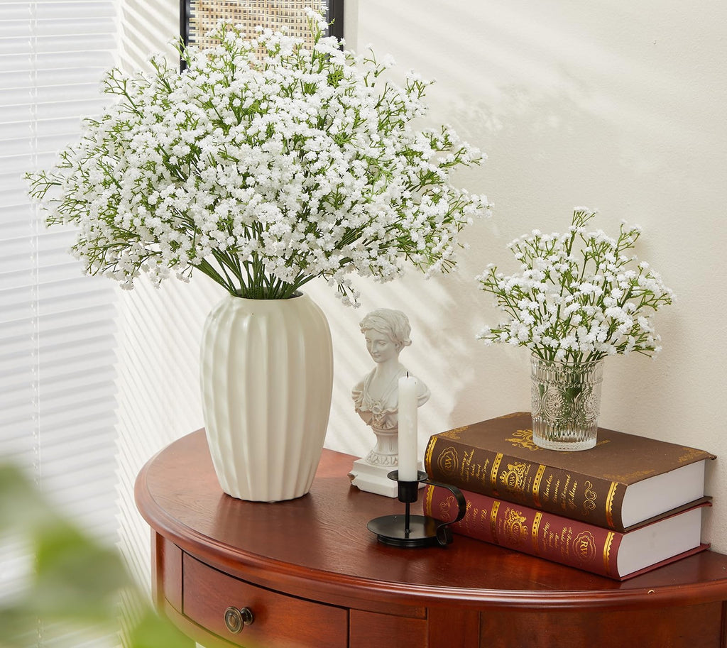 Two bouquets of white artificial baby's breath flowers in vases on a wooden table with books and a bust.