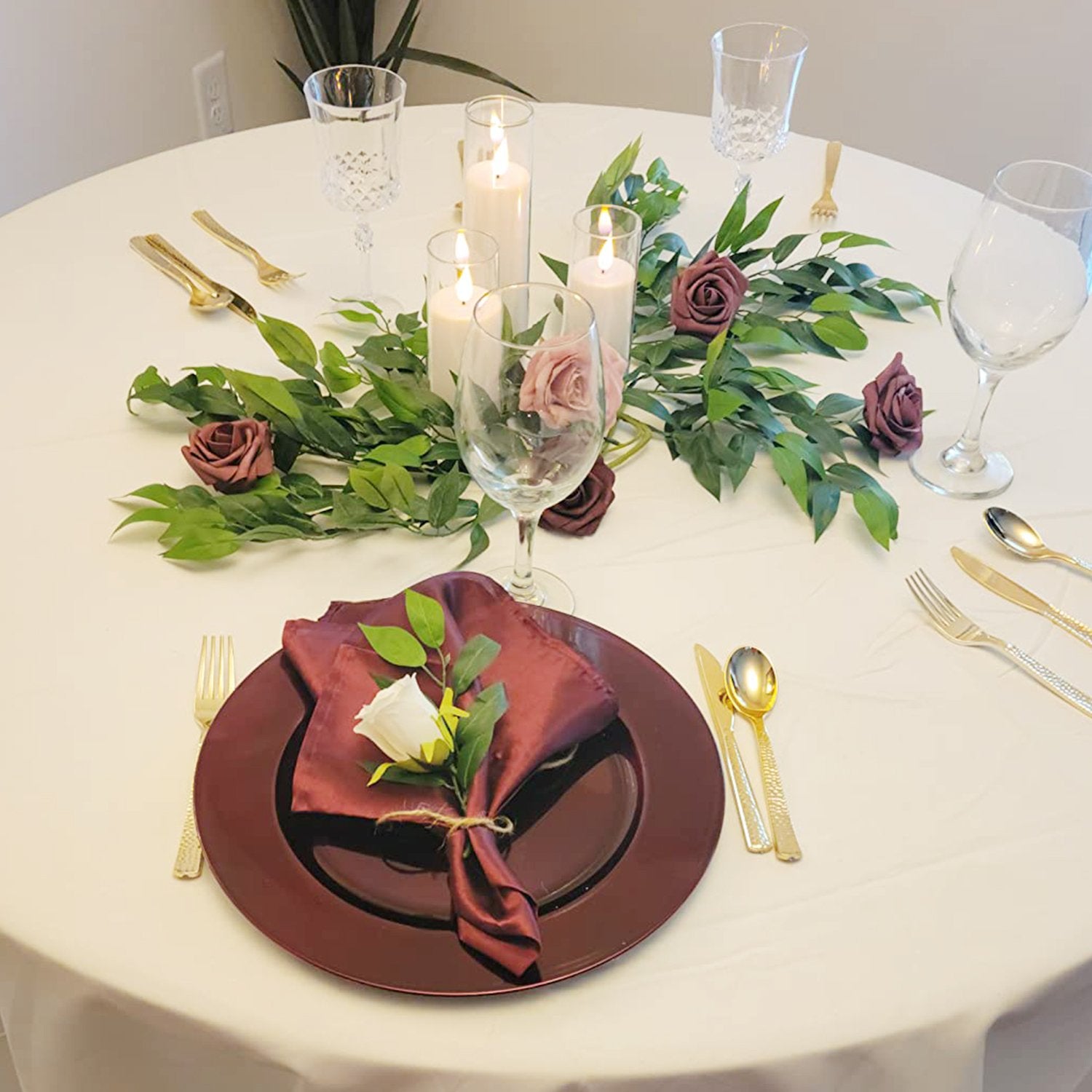 Table setting with burgundy charger plate, napkin, and gold cutlery, adorned with greenery and roses, surrounded by candles and wine glasses.