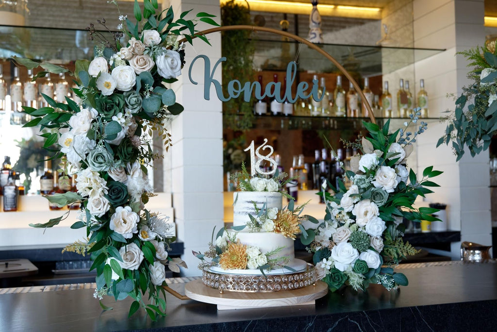 Two-tier cake with 'Konalei' and '18' toppers, surrounded by floral arrangements on a bar counter.