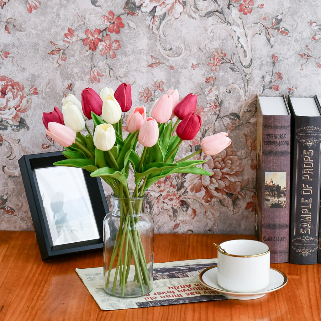 Artificial tulips in shades of pink, white, and red arranged in a glass vase on a wooden table with books and a teacup.