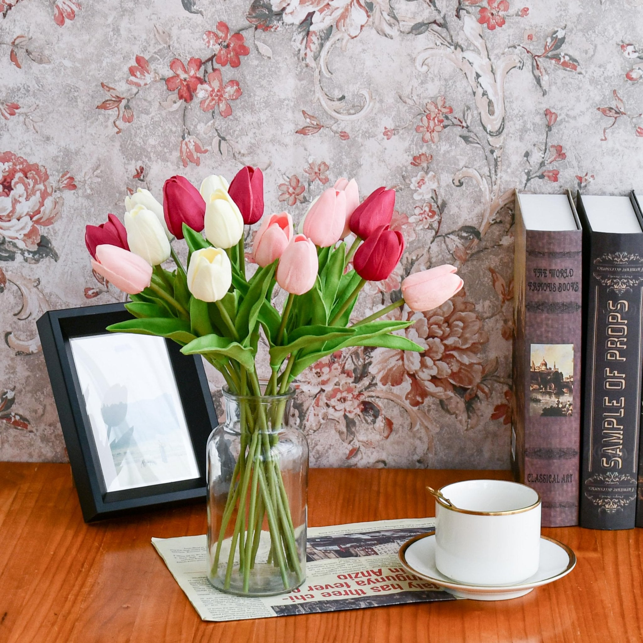 Artificial tulips in shades of pink, white, and red arranged in a glass vase on a wooden table with books and a teacup.