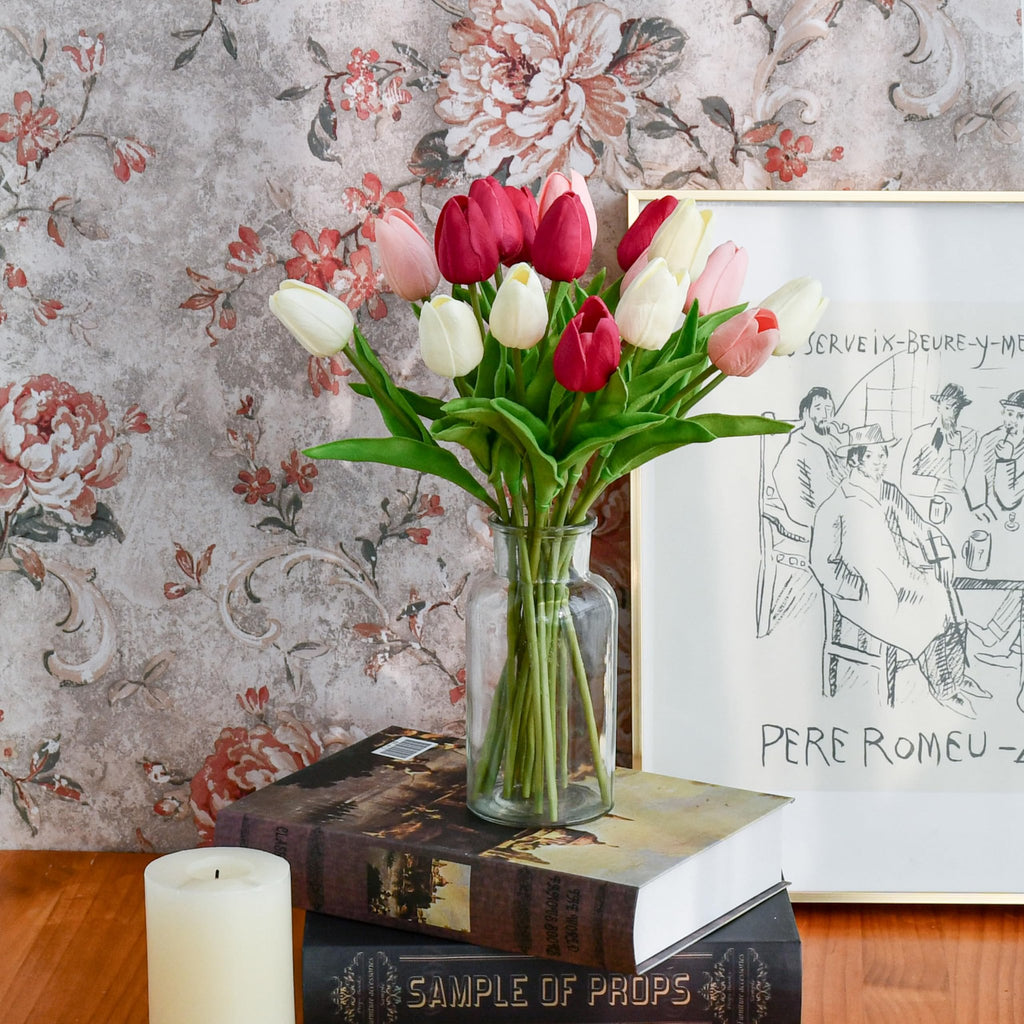 Artificial tulips in shades of pink, red, and white in a glass vase on stacked books, with a candle and framed art.